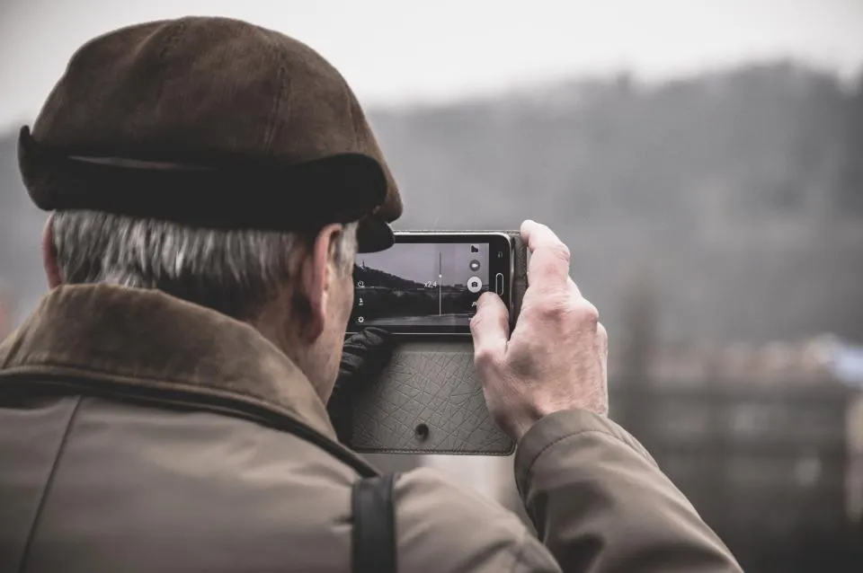Older man in a cap taking a photo with a phone outdoors